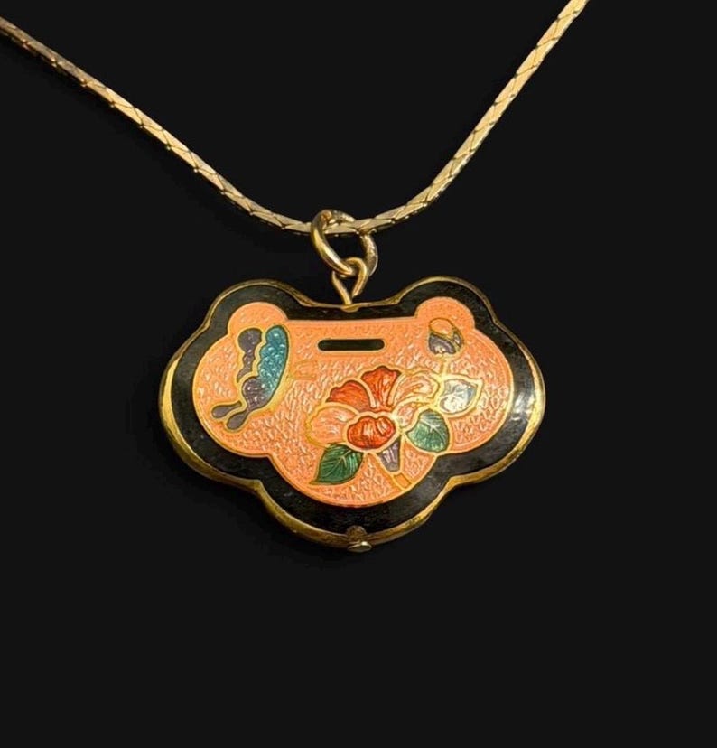 May include: A gold-toned necklace with a pendant featuring a floral design. The pendant has a peach-colored background with a black border and a butterfly and flower design. The necklace is on a black background.