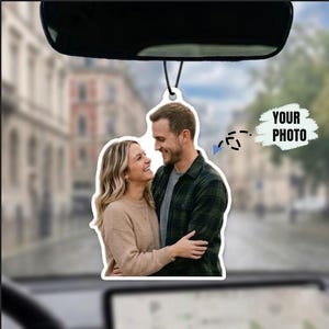 May include: A custom car air freshener hanging from a rearview mirror, featuring a printed photograph of a smiling couple embracing. The person on the left wears a beige sweater, and the person on the right wears a green plaid shirt. Text reads YOUR PHOTO.
