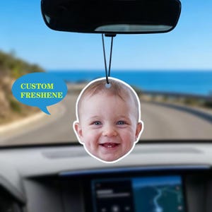 Peut inclure: Un désodorisant de voiture personnalisé avec le visage souriant d'un bébé. Le désodorisant est rond avec un bord blanc et est suspendu à une ficelle noire. Le texte "CUSTOM FRESHENE" est dans une bulle bleue.