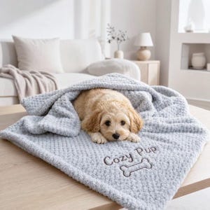 Pode incluir: Uma manta de pelúcia cinza-claro texturizada com o texto bordado "Cozy Pup" acima de um ícone em forma de osso. Um pequeno filhote de cor clara com orelhas caídas está deitado, parcialmente coberto pelo tecido macio e com covinhas.