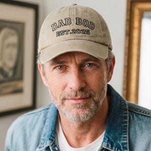 Peut inclure: Casquette de baseball beige avec l'inscription "DAD BOD EST. 2025" en noir. La casquette est portée par une personne portant une veste en jean et un t-shirt blanc.