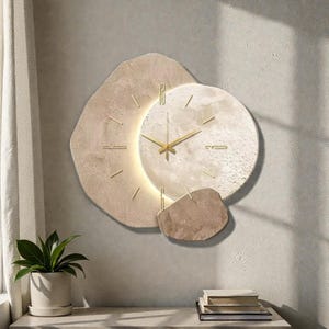 Op de afbeelding: Een moderne wandklok met een uniek ontwerp. De klok heeft een lichtbeige, getextureerde wijzerplaat met goudkleurige wijzers en markeringen. De klok is omgeven door onregelmatig gevormde beige elementen, wat een eigentijdse uitstraling creëert. De klok is aan een lichtgrijze muur bevestigd.