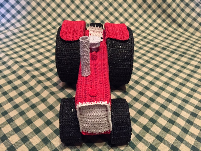 Pu&ograve; includere: Un trattore giocattolo in crochet rosso e nero con un tubo di scarico in crochet bianco.