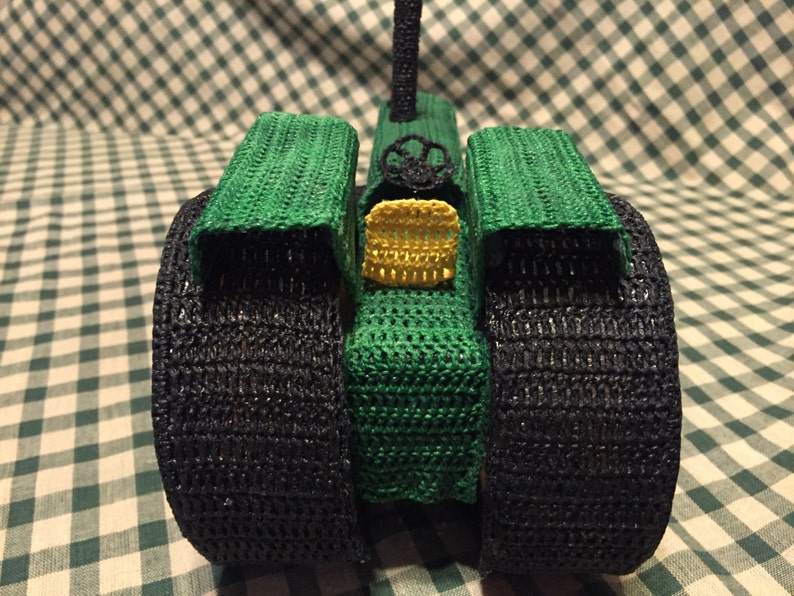 May include: A green and black crocheted toy tractor with large wheels. The tractor has a yellow crocheted detail on the front.