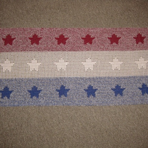 Puede incluir: Una alfombra de crochet roja, blanca y azul con estrellas. La alfombra tiene tres filas de estrellas, con estrellas rojas en la fila superior, estrellas blancas en la fila del medio y estrellas azules en la fila inferior.