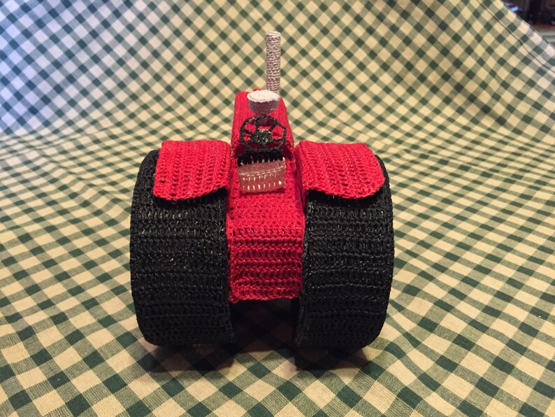 Pu&ograve; includere: Un trattore giocattolo in crochet rosso con ruote nere. Il trattore ha un piccolo camino in crochet bianco in cima.