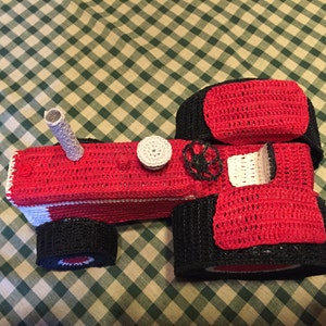 Pu&ograve; includere: Un trattore giocattolo in crochet rosso e nero con un tetto in crochet bianco e un tubo di scarico in crochet bianco.