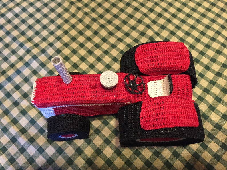 Pu&ograve; includere: Un trattore giocattolo in crochet rosso e nero con un tetto in crochet bianco e un tubo di scarico in crochet bianco.