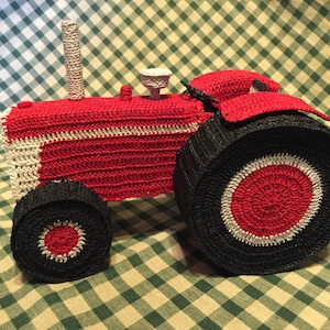 Pu&ograve; includere: Un trattore giocattolo in crochet rosso e nero con accenti bianchi. Il trattore ha una grande ruota posteriore e una ruota anteriore pi&ugrave; piccola.
