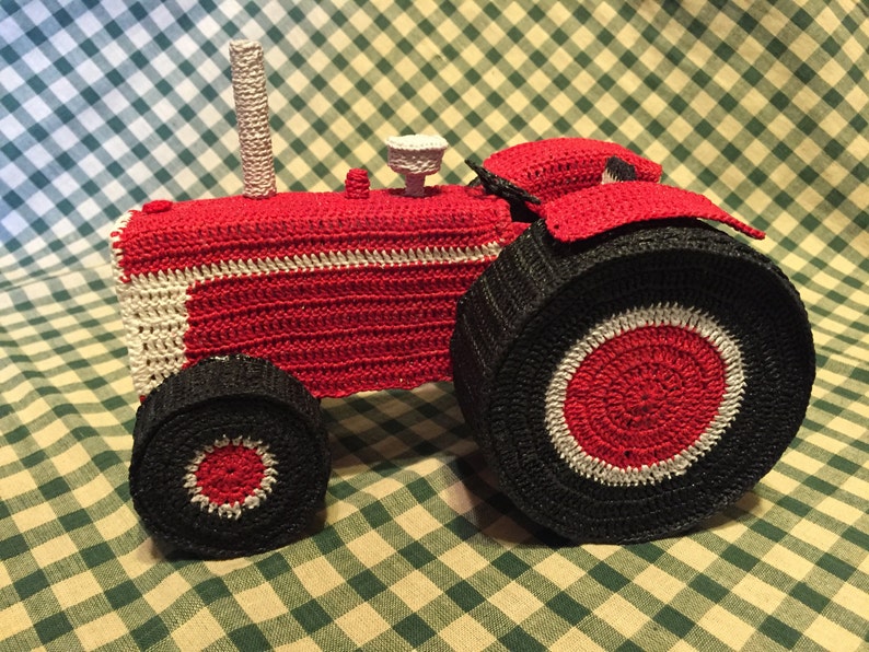 Pu&ograve; includere: Un trattore giocattolo in crochet rosso e nero con accenti bianchi. Il trattore ha una grande ruota posteriore e una ruota anteriore pi&ugrave; piccola.