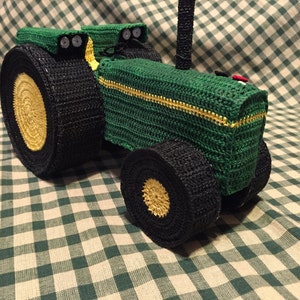 May include: A green crocheted toy tractor with black wheels and yellow accents. The tractor has a black exhaust pipe and a red light on the back.