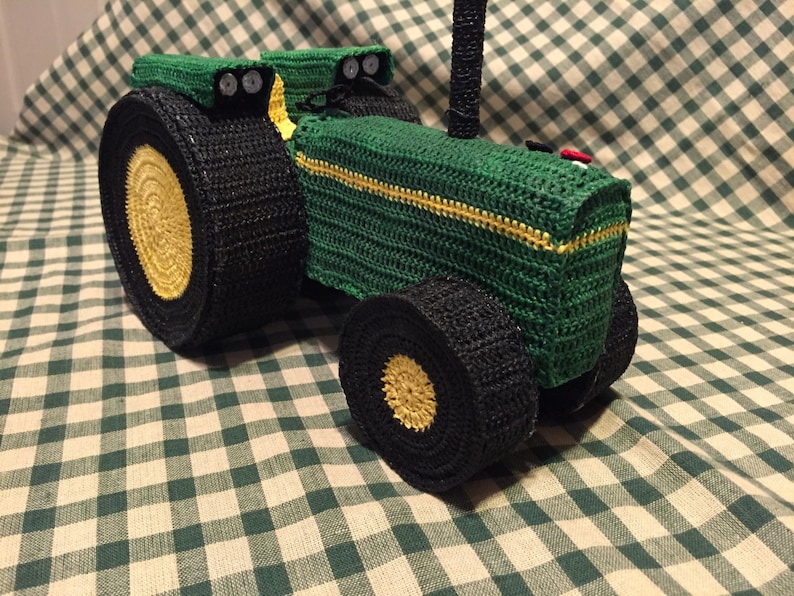 May include: A green crocheted toy tractor with black wheels and yellow accents. The tractor has a black exhaust pipe and a red light on the back.