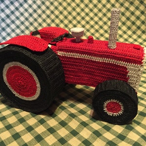 Pu&ograve; includere: Un trattore giocattolo in crochet rosso e nero con una striscia bianca. Il trattore ha una grande ruota anteriore e una ruota posteriore pi&ugrave; piccola.