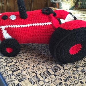 May include: A red and black crocheted tractor toy. The tractor has a white stripe around the middle and black wheels.