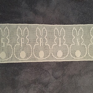 Op de afbeelding: Witte gehaakte kant met een herhalend patroon van gestileerde konijnen. De kant is gemaakt van fijn, delicaat garen en heeft een gegolfde rand.