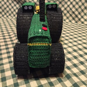 May include: A green and black crocheted toy tractor with large black wheels. The tractor has a red button on the front.