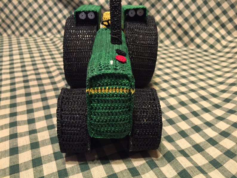 May include: A green and black crocheted toy tractor with large black wheels. The tractor has a red button on the front.