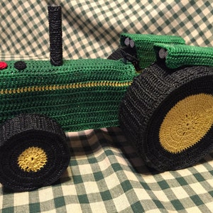 May include: Green crocheted toy tractor with black wheels and yellow tire treads. The tractor has a black exhaust pipe and a black seat.