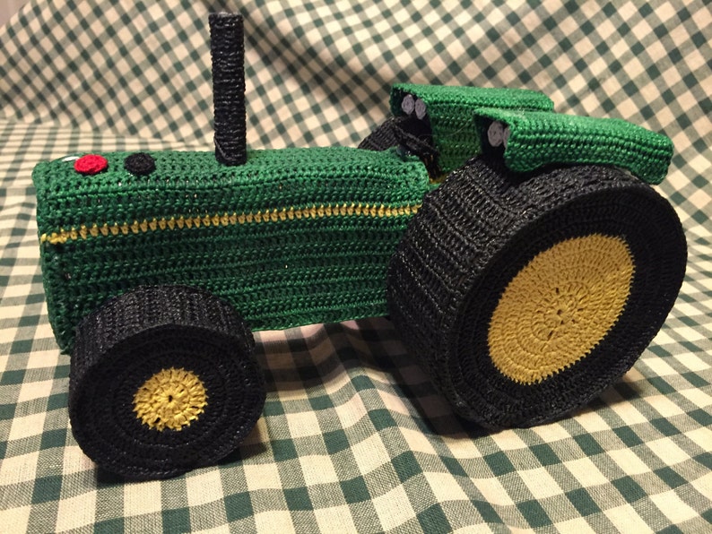May include: Green crocheted toy tractor with black wheels and yellow tire treads. The tractor has a black exhaust pipe and a black seat.