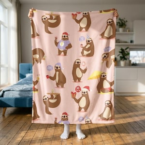 Könnte beinhalten: Eine hellrosa Fleecedecke mit einem sich wiederholenden Muster aus braunen Faultieren bei verschiedenen Aktivitäten, darunter das Halten von Geschenken, das Tragen von Partyhüten, Schlafen und Kaffeetrinken, mit Sprechblasen wie Good Morning und Ho Ho Ho.