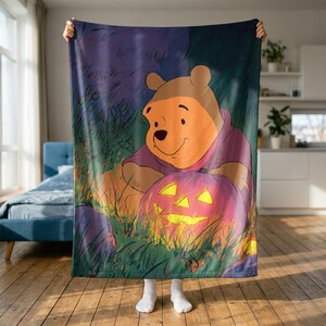 Puede incluir: Una manta de forro polar rectangular con una ilustración impresa de un oso de color marrón claro con una camiseta morada, sentado en un campo de hierba oscura junto a una calabaza naranja brillante con cara tallada, bajo un cielo morado.