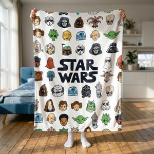 Könnte beinhalten: Eine weiße Fleecedecke mit einem sich wiederholenden Muster aus verschiedenen Star Wars-Charakterköpfen und Helmen im Cartoon-Stil, wobei der fettgedruckte schwarze Text STAR WARS in der Mitte des Designs zwischen den Symbolen steht.