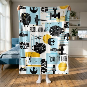 Op de afbeelding: Een witte fleece deken met een herhalend Star Wars-patroon met zwarte silhouetten van X-wing starfighters, Millennium Falcon schepen en Rebel Alliance logo's, voorzien van lichtblauwe en gele rechthoekige accenten en tekst.