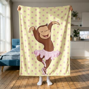 May include: A soft fleece blanket featuring a light yellow background with a repeating pattern of pink polka dots. In the centre, a brown monkey character is depicted wearing a light pink ballet tutu and matching ballet slippers while dancing.