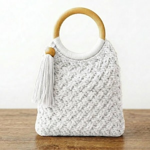 May include: A light gray crocheted handbag with a wooden handle. The bag features a textured stitch pattern and a matching tassel. The handle is a large, circular wooden ring. The bag is sitting on a wooden surface.