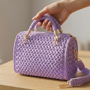 May include: A lavender handbag featuring a woven cord texture, a cylindrical shape, and gold-toned metal hardware. The bag includes a sturdy top handle and a detachable shoulder strap, both crafted from the same lavender cord material.
