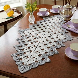 Puede incluir: Camino de mesa de ganchillo calado hecho a mano en blanco y gris, con borde festoneado. El camino de mesa está colocado sobre una mesa de madera, junto a tazas de té, una tetera y un jarrón con flores. Se ve una madeja de lana amarilla.