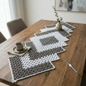 May include: A handmade crochet table runner in a zig-zag pattern, featuring alternating sections of white and gray. A small ceramic vase with a sprig of greenery and a coffee cup with a saucer are on the wooden table. A crochet hook rests on the table.