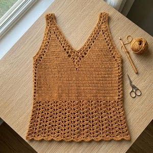 May include: A handmade, mustard-colored crochet top with a V-neck design. The top features a solid stitch pattern on the upper portion and a decorative openwork pattern on the lower portion. A ball of yarn, crochet hook, and scissors are also visible.