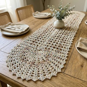 May include: A cream-colored, open-weave crochet table runner with a scalloped edge. The runner is placed on a wooden table, with place settings and a small vase with greenery. The runner's design features small flecks of color.