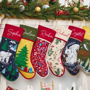 Puede incluir: Seis calcetines navideños personalizados colgados en fila. Cada calcetín presenta un patrón único que incluye un oso polar, un árbol de Navidad, motivos florales, hojas de acebo, una ardilla y un alce, con nombres bordados en los puños.