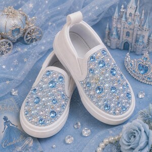 Puede incluir: Un par de zapatillas blancas sin cordones con una densa decoración de cristales azul claro y transparentes en el empeine. Los zapatos tienen una suela de goma blanca gruesa y una lengüeta en el talón, con un diseño brillante en la parte frontal.