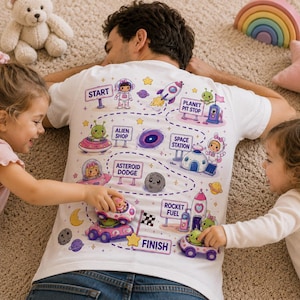 Puede incluir: Camiseta blanca con un diseño de mapa colorido con temática espacial. El mapa incluye ilustraciones de extraterrestres, cohetes, planetas y estaciones espaciales. La camiseta está siendo manipulada por dos niños.