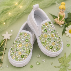 Könnte beinhalten: Ein Paar weiße Slipper mit einer dichten Verzierung aus grünen, gelben und klaren Kristallen auf der Vorderkappe und den Seitenteilen. Die Schuhe haben eine saubere weiße Gummisohle und eine Zuglasche an der Ferse mit funkelnder Textur.