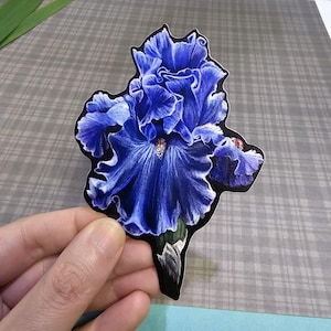 May include: A sticker featuring a detailed illustration of a vibrant blue iris flower. The flower has ruffled petals and a dark outline. The sticker is held against a gray plaid background.