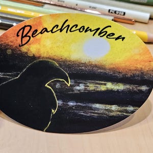 May include: A round sticker with a black silhouette of a bird watching a sunset over the ocean. The sticker has the text "Beachcomber" written in black cursive.