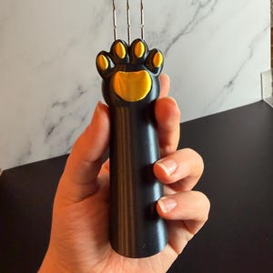 Könnte beinhalten: Ein schwarz-gelbes Werkzeug in Form einer Katzenpfote mit drei Metallnadeln. Das Werkzeug wird in einer Hand gehalten. Die Pfotenballen sind gelb. Der Hintergrund ist weißer und grauer Marmor.