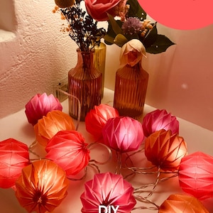 Può includere: Una stringa di lanterne di carta origami rosa e arancioni, illuminate con luce calda. Le luci sono disposte su una superficie bianca, con vasi di fiori sullo sfondo. Il testo sull'immagine recita "DIY Guirlande lumineuse Origami".