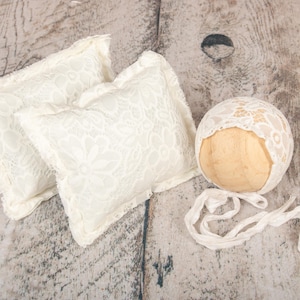 May include: Cream-colored newborn photography props, including two square pillows and a lace bonnet. The pillows feature a floral lace pattern. The bonnet is designed to fit a newborn's head and has a matching lace design.