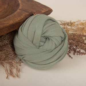 May include: A sage green stretchy fabric wrap for newborn photography. The wrap is rolled up and is sitting on a rustic wooden bowl and burlap fabric.