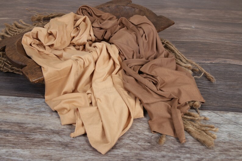 May include: Two fabric swaddles in beige and brown colors, arranged in a wooden bowl on a woodgrain background.