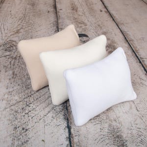 May include: Three rectangular pillows in different shades of white, beige, and cream. The pillows are arranged in a row on a wooden surface.