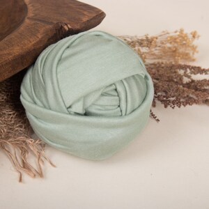 May include: A sage green stretchy fabric wrap for newborn photography. The fabric is rolled up into a ball shape.