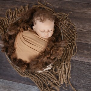 May include: A newborn baby doll is wrapped in a tan swaddle and placed in a brown woven basket. The basket is on a dark brown wooden surface.