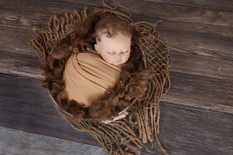 May include: A newborn baby doll is wrapped in a tan swaddle and placed in a brown woven basket. The basket is on a dark brown wooden surface.
