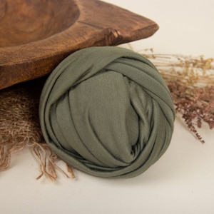 May include: A tightly rolled green fabric swaddle, perfect for newborn photography. The swaddle is lying on a rustic wooden bowl and burlap fabric.
